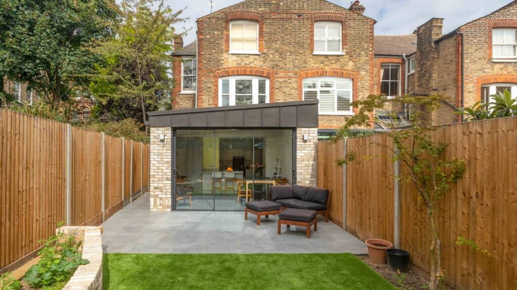 Exterior view of a London house featuring a kitchen extension with black cladding and wood paneling, large black-framed glass doors opening to the garden, and timber slats allowing natural light to filter through.