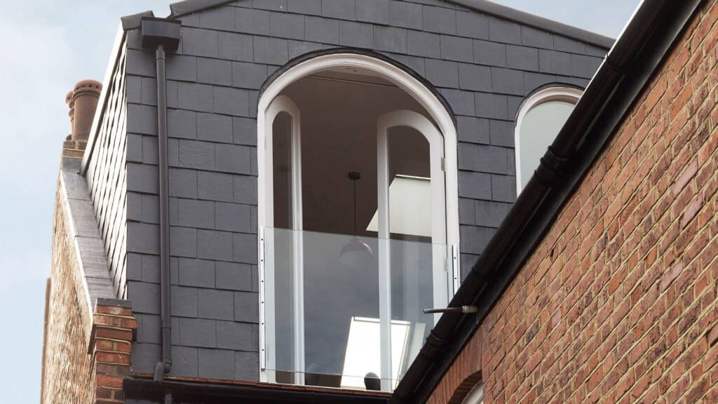 An arched loft window with a small balcony against black slate and London brickwork.