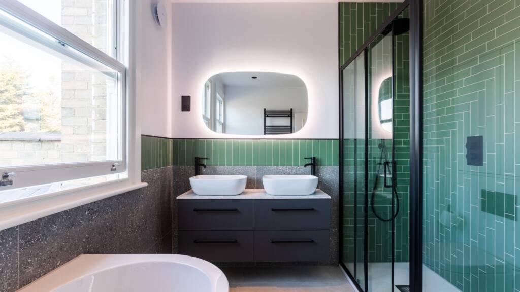 Modern bathroom featuring a floating wall-hung vanity with dual sinks, matte black taps and showerheads, and bold green geometric tiles with terrazzo accents.